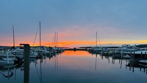 Eine Marina mit Booten an hölzernen Stegen, ruhigem Wasser, das die Szene spiegelt, und einem Sonnenuntergang im Hintergrund.
