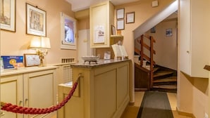 A hotel lobby with a reception desk, framed pictures on the walls, a staircase, and a red rope barrier. - Casa Miramonte (Bologna)