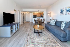 A living room with a flat-screen TV, a gray sofa, a patterned rug, and a dining area with wooden cabinets and a chandelier. - Updated 5th Floor Condo w/ Fun Amenities at The Palms of Destin! 💚🥥🌴 (Destin)