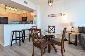 A modern kitchen with wooden cabinets, a dining area with a glass table and wooden chairs, and a cozy seating area with a sofa and lamp. - Updated 5th Floor Condo w/ Fun Amenities at The Palms of Destin! 💚🥥🌴 (Destin)