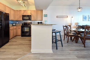 A modern kitchen with wooden cabinets, a central island, and a dining area with chairs and a table. - Updated 5th Floor Condo w/ Fun Amenities at The Palms of Destin! 💚🥥🌴 (Destin)