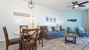 Ein modernes Wohnzimmer mit Esstisch, Stühlen, einer Couch und einem Couchtisch. Es gibt einen Deckenventilator, Wandbilder und einen Teppich auf dem Boden.