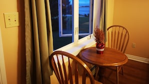 A room with a wooden table and two chairs, a window with curtains, a small plant on the windowsill, and a wall-mounted hanger.