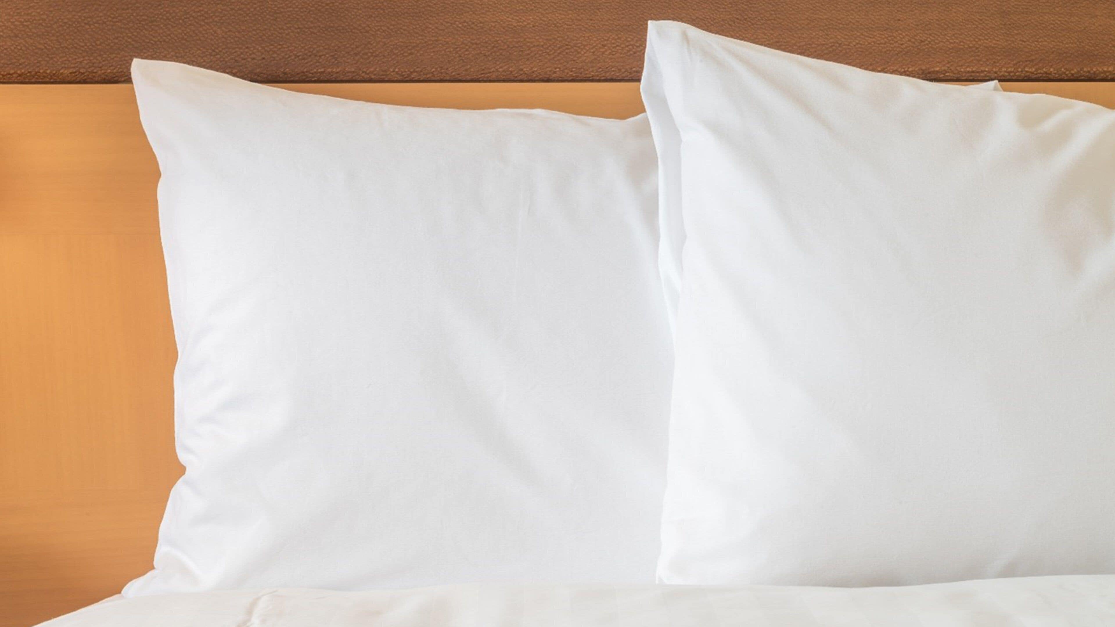 Una cama con sábanas y dos almohadas blancas, apoyadas contra un cabecero marrón.
