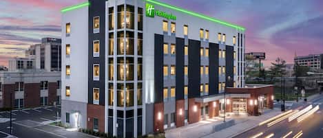 A Holiday Inn hotel with a modern design, featuring a prominent green sign, multiple windows, and a well-lit entrance.