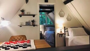 Een gezellig zolderkamer met een bed, een klein tafeltje en een schaakbord. Er staan planken met planten en een raam met uitzicht op bomen.