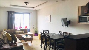 A modern living room with a sofa, dining table, and chairs. There is a TV mounted on the wall and a window with curtains. - Residence Hivernage Founty 5429 (Agadir)