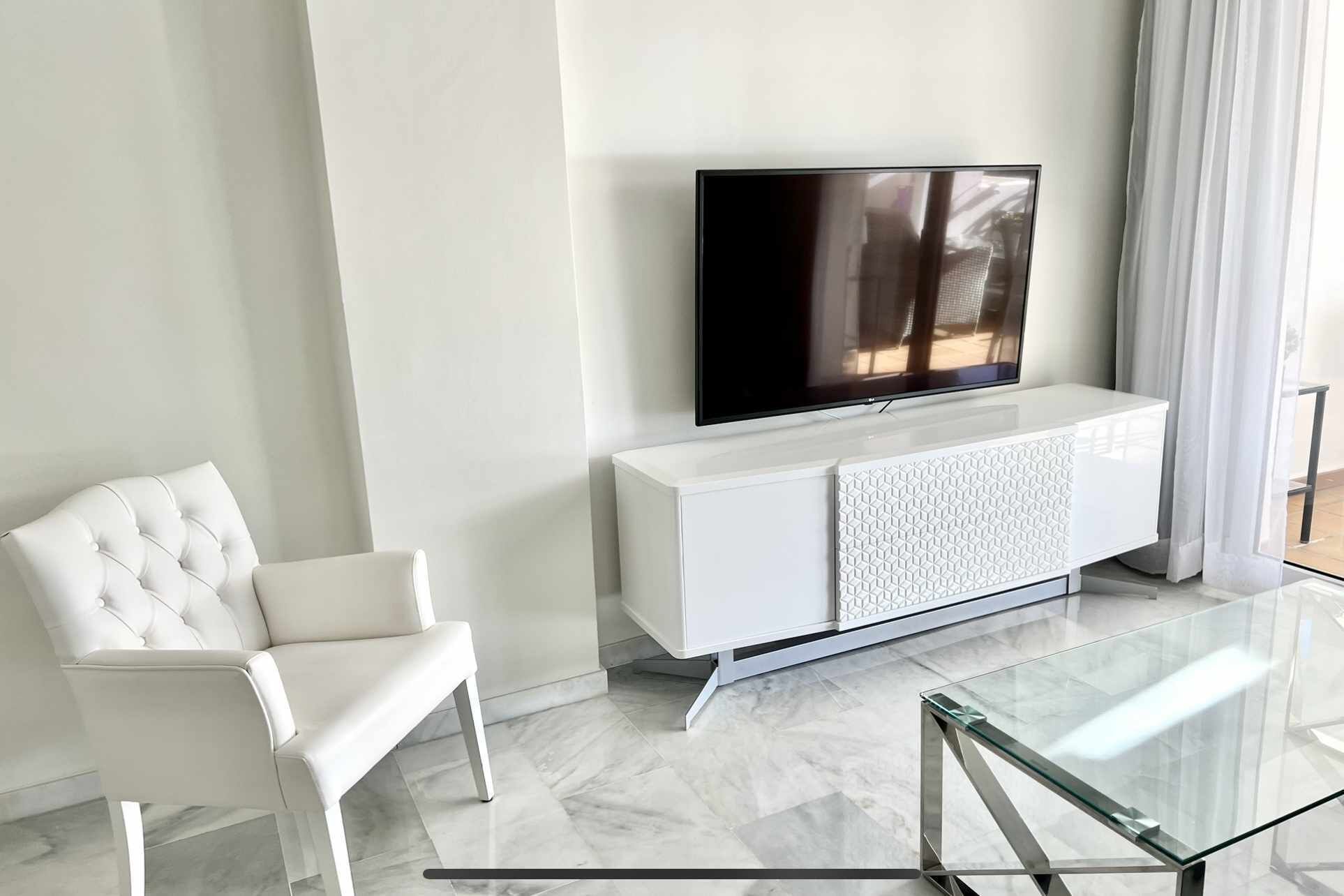 Een moderne woonkamer met een flatscreen televisie bevestigd aan een witte kast, een gestoffeerde witte fauteuil en een glazen salontafel.