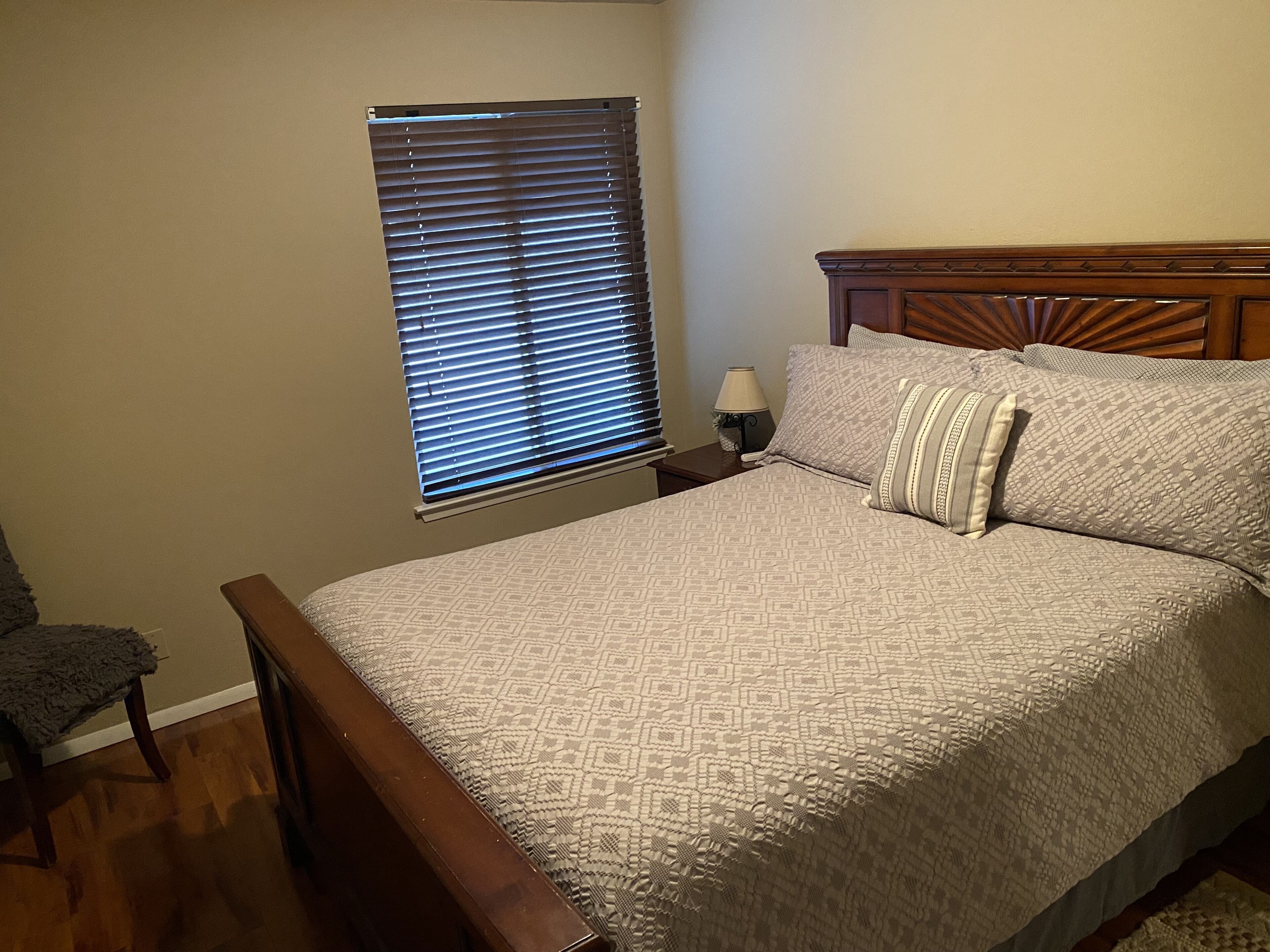 A bedroom with a bed, a nightstand, a lamp, a window with blinds, and a chair.