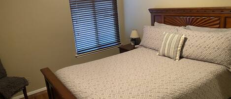 A bedroom with a bed, a nightstand, a lamp, a window with blinds, and a chair.