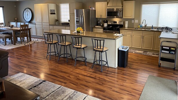 A modern kitchen with a central island, bar stools, stainless steel appliances, and a dining area with a clock and bar stools. - Casa De La Roca (Albuquerque)