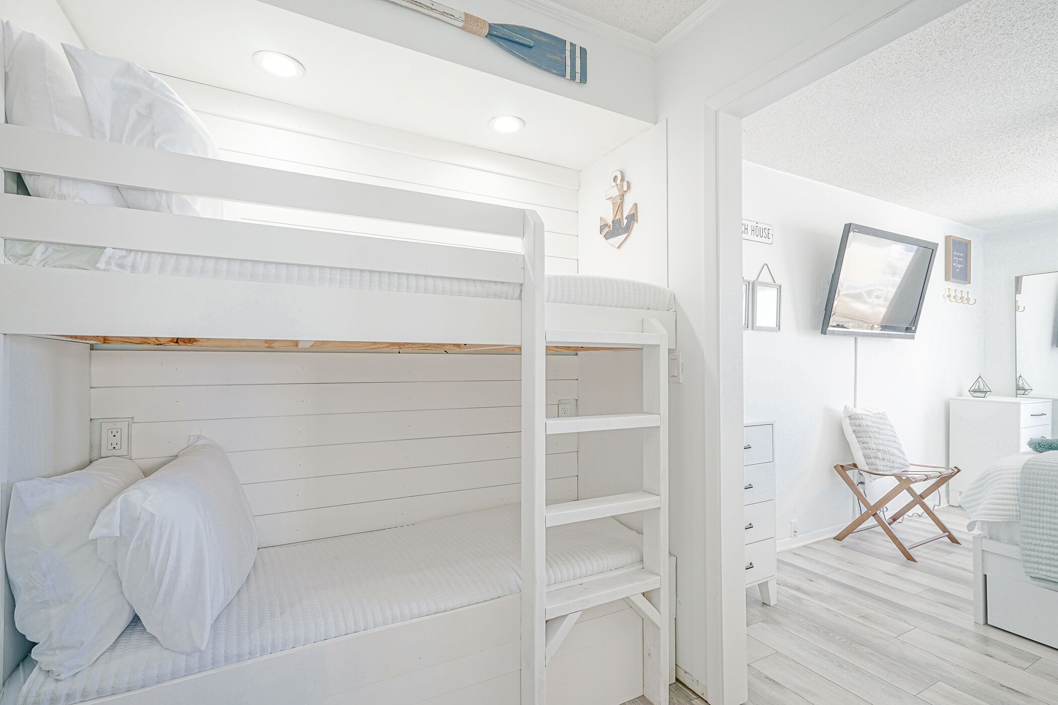 Een slaapkamer met stapelbed, een witte kleurstelling, een aan de muur bevestigde televisie en een stoel met kussen.