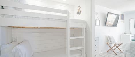Een slaapkamer met stapelbed, een witte kleurstelling, een aan de muur bevestigde televisie en een stoel met kussen.