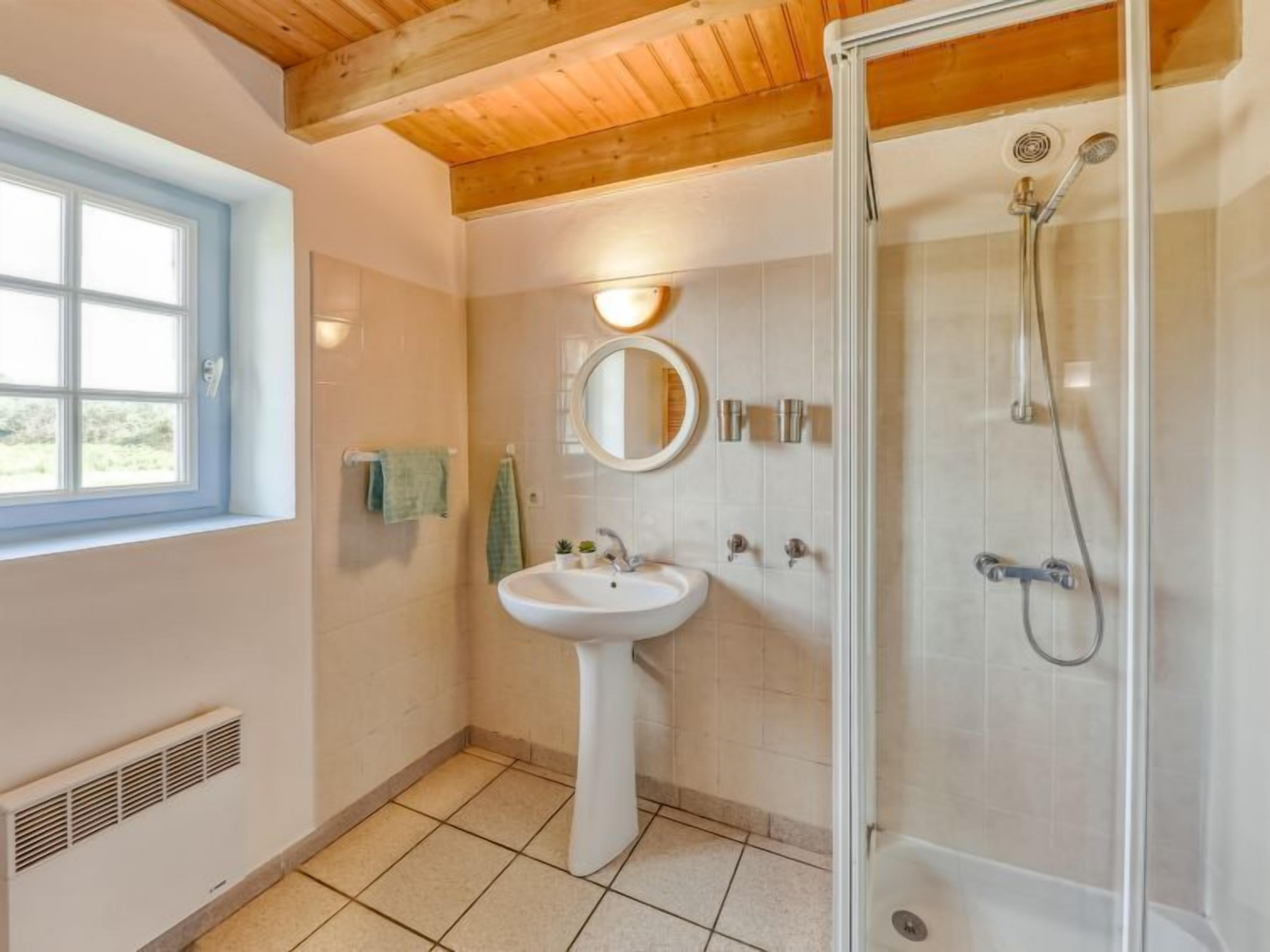 A bathroom with a pedestal sink, a round mirror, a glass-enclosed shower, and a window providing natural light.