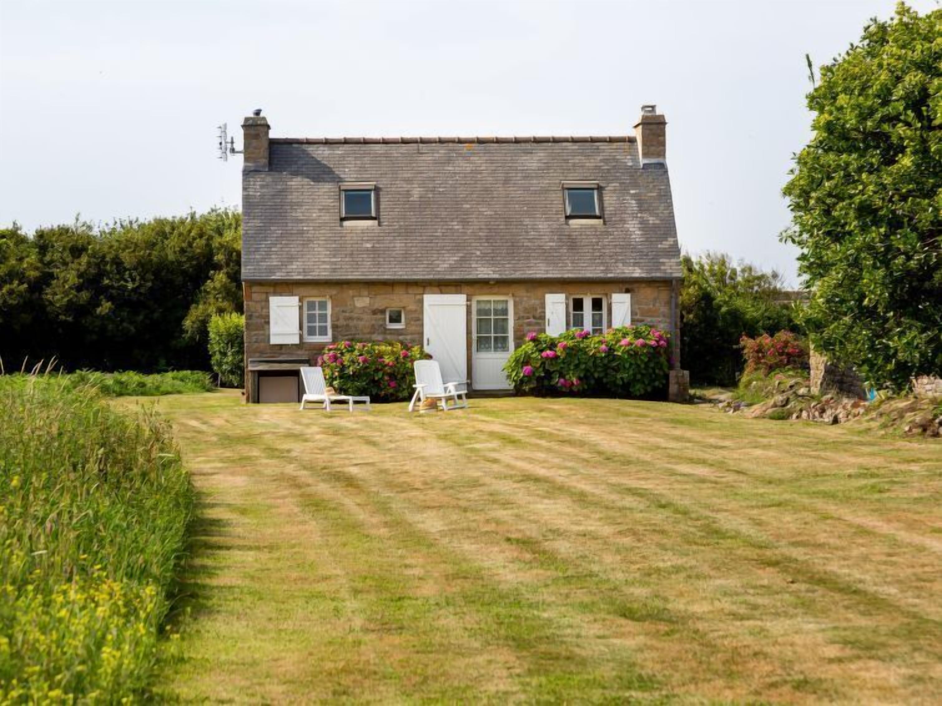Une maison en pierre avec un toit en pente, deux fenêtres, une porte et un petit garage. Il y a deux chaises blanches dans le jardin devant la maison.