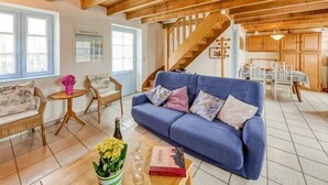 A cozy living room with a blue sofa, wooden ceiling beams, a staircase, and a dining area with white chairs. - Ker Kébec by Interhome (Crozon-Morgat)