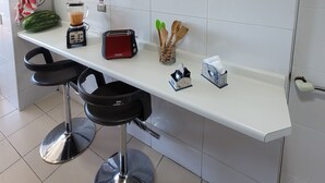 A kitchen with a white countertop, a blender, a toaster, a small refrigerator, and a fruit. There are two black bar stools in front of the counter.