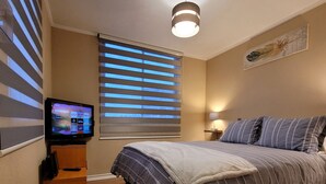 A bedroom with a bed, a TV, a window with blinds, a lamp, and a ceiling light.