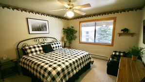 A bedroom with a bed, a ceiling fan, a window with blinds, a framed picture on the wall, and a small shelf with a plant.