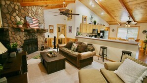 A cozy living room with a stone fireplace, leather sofa, and a kitchen area with wooden cabinets and a bar.