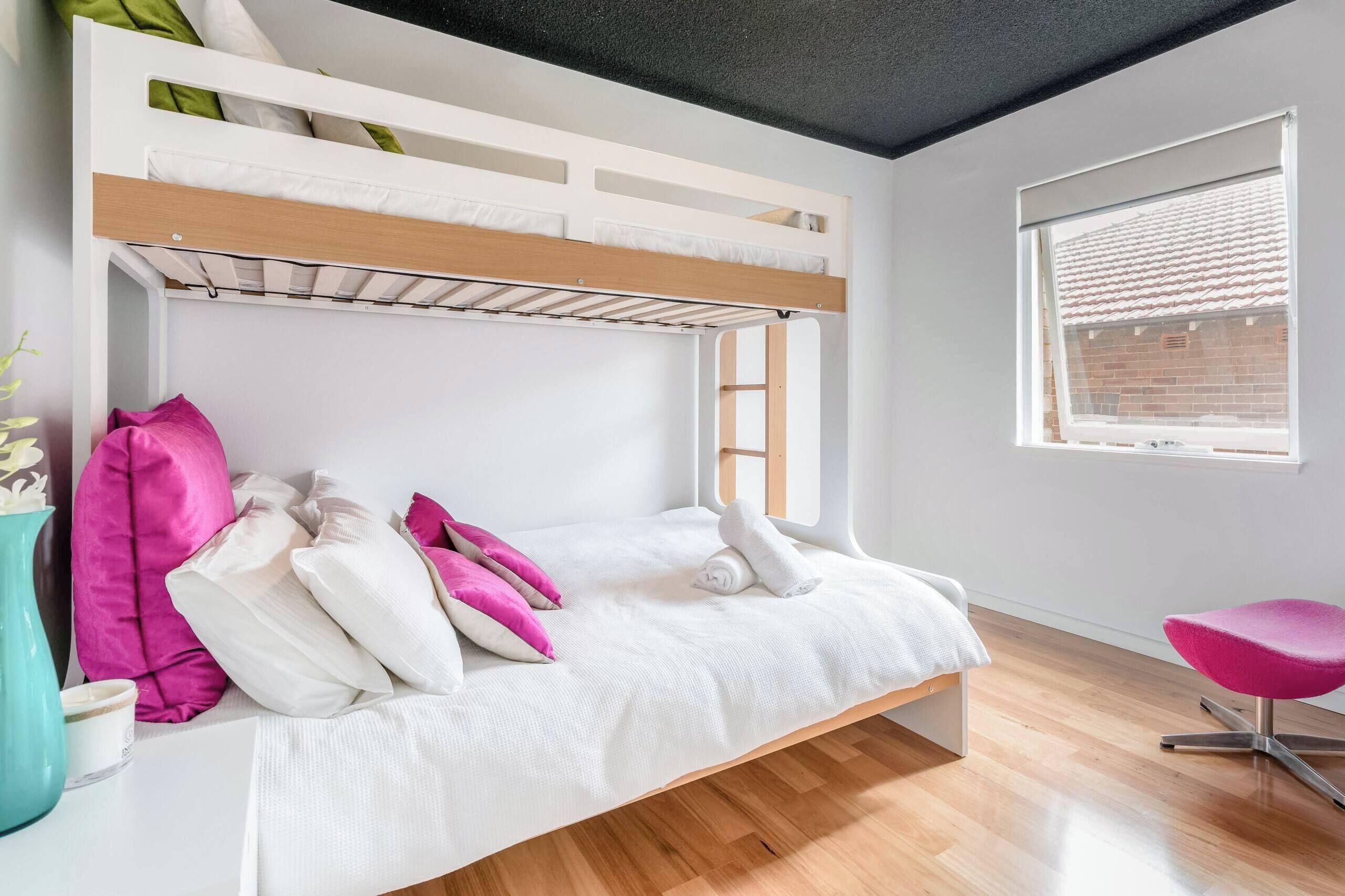 Uma cama beliche com colchão branco e almofadas cor-de-rosa, um vaso verde-água e uma cadeira rosa num quarto com piso de madeira e uma janela com persianas.
