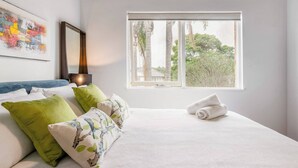 Un lit bien fait, avec une literie blanche et des coussins verts, une grande fenêtre donnant sur des arbres et un tableau abstrait aux couleurs vives accroché au mur.