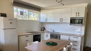 Cozinha moderna com armários brancos, eletrodomésticos de aço inoxidável, mesa de jantar de madeira e cadeiras brancas.