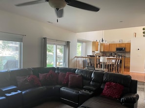 A spacious living room with a black leather sectional sofa, a ceiling fan, and a view of the kitchen and dining area through large windows.