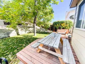 Una terraza de madera con una mesa de picnic, rodeada por un césped y árboles.
