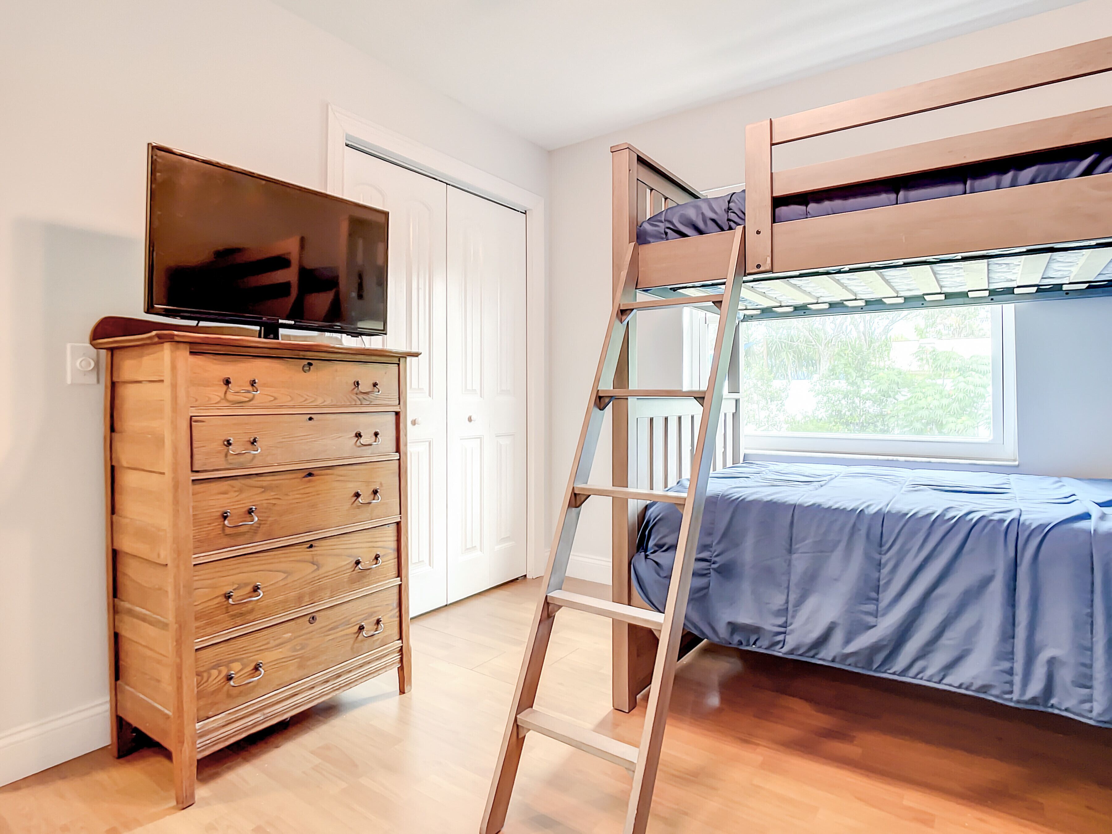 A room with a bunk bed, a wooden dresser with drawers, a television on top, and a window with a view of greenery.