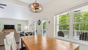 A spacious living room with a wooden dining table, a sofa, and a TV. There are large windows with a view of trees. - Livin' the Dream in Fish Creek! (Fish Creek)