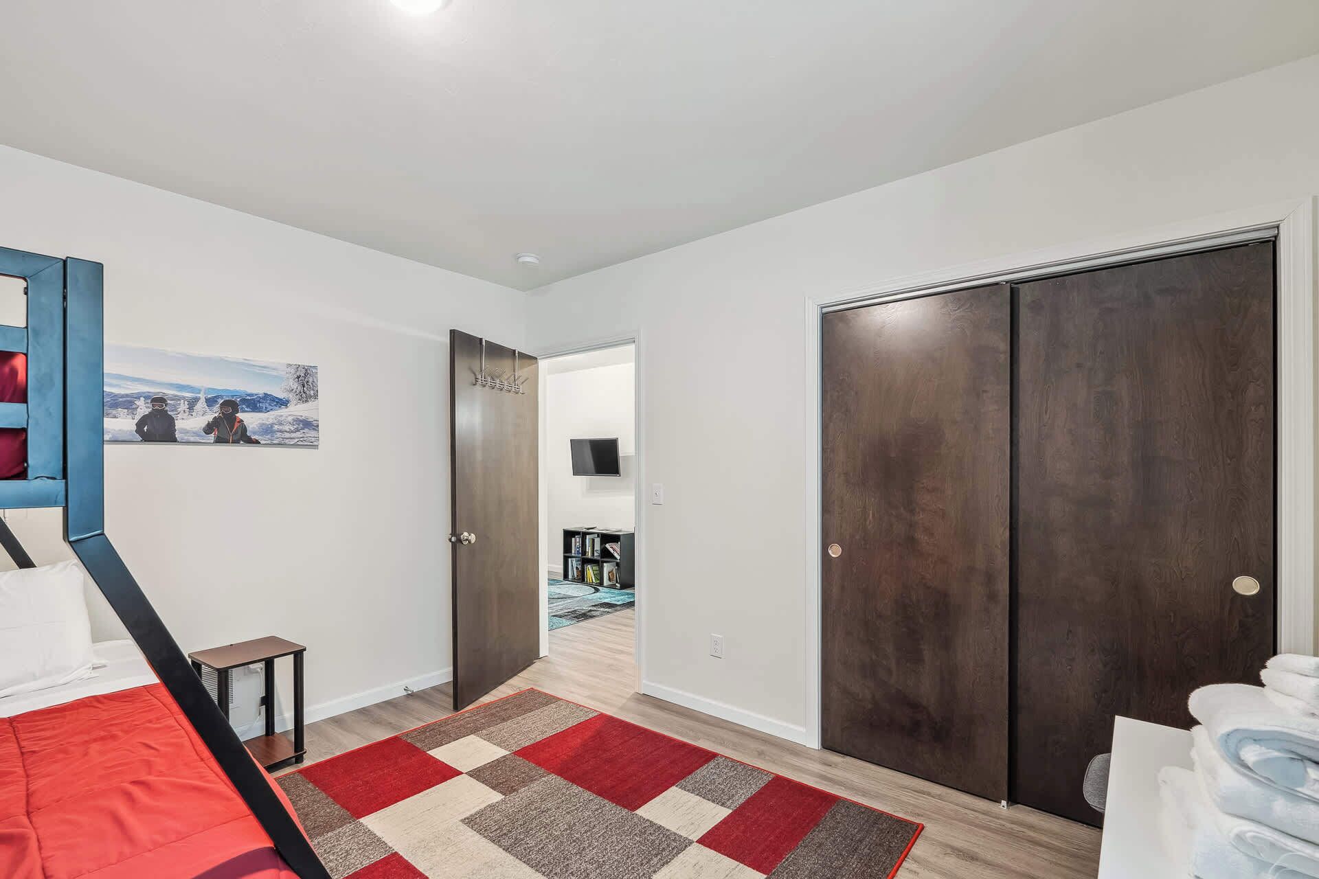 A bedroom with a bed, a red and beige checkered rug, a dark wooden wardrobe, and a hallway leading to a bathroom.