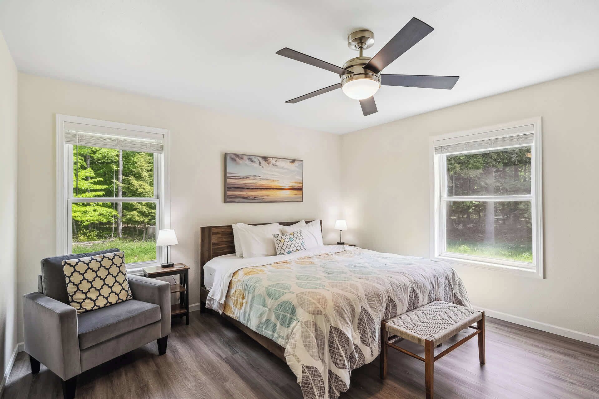 A bedroom with a bed, a chair, a desk, a ceiling fan, and two windows.