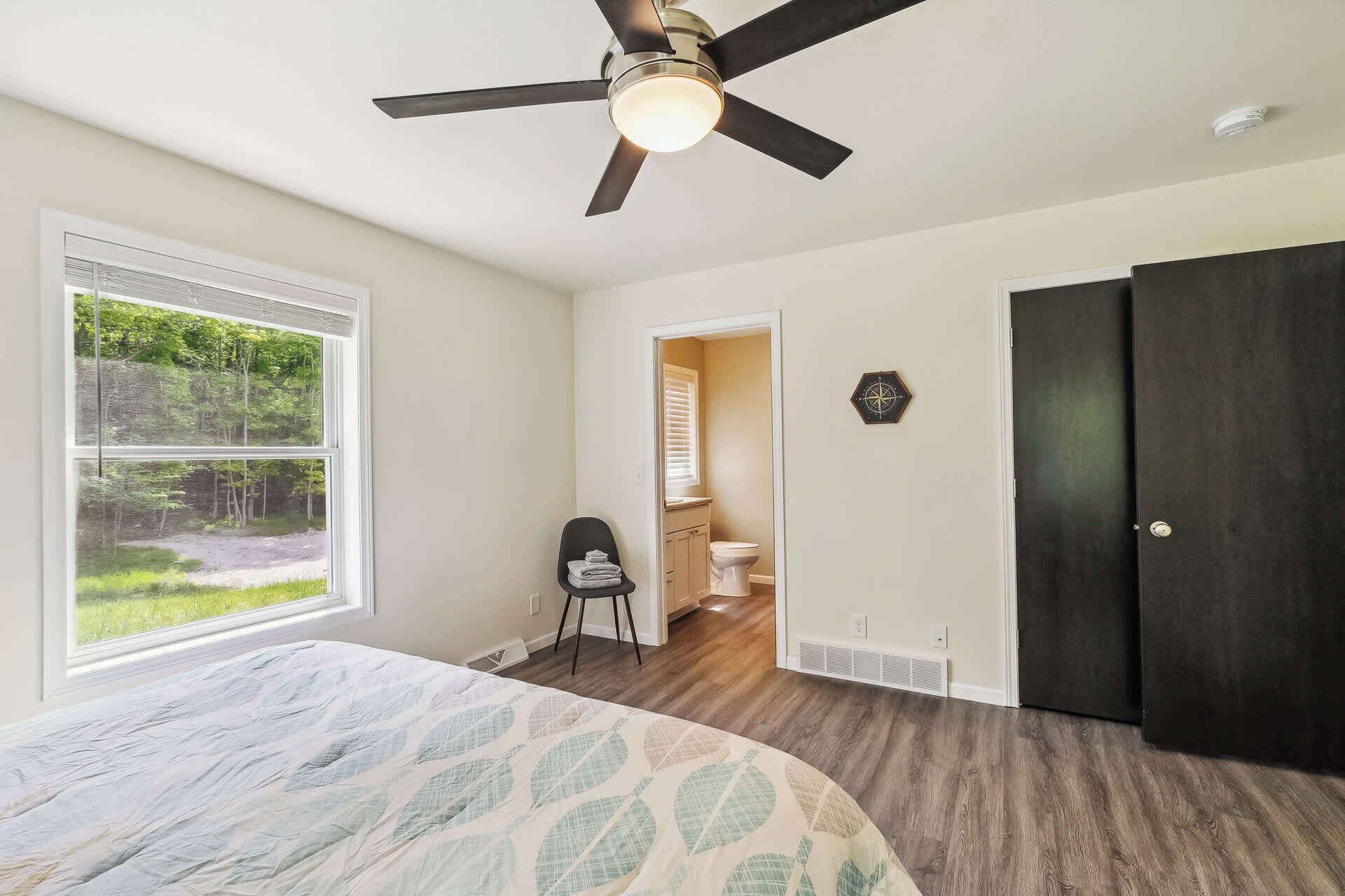 A bedroom with a bed, a ceiling fan, a window with a view of greenery, and a door leading to a bathroom.