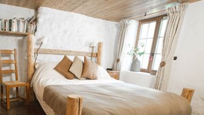 A bedroom with a wooden headboard, a large bed with a beige blanket, a wooden chair, and a window with curtains. - Savoyard Farmhouse - Sleeps 14, Garden & Parking (Araches-la-Frasse)