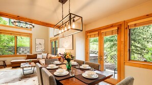 A dining area with a wooden table set for four, surrounded by chairs, with a view of greenery through large windows. - Year-Round Fun - Central Location, Private Hot Tub & 5-Min Walk to Free Shuttle! (Park City)