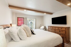 Un dormitorio con una cama grande, cabecera de madera, televisor montado en la pared y una ventana con cortinas.