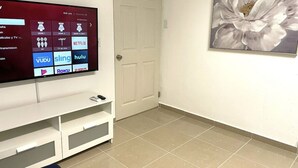 A modern living room with a flat-screen TV mounted on a white entertainment unit, a door, and a large floral painting on the wall. - California Apartments (Yabucoa)