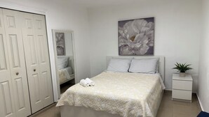 A bedroom with a bed, a nightstand, a potted plant, and a door. - California Apartments (Yabucoa)
