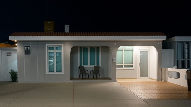 A modern house with a tiled roof, a covered patio with chairs, and a door leading to the interior. - California Apartments (Yabucoa)