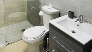 A modern bathroom with a glass-enclosed shower, a toilet, a sink, and a mirror. - California Apartments (Yabucoa)