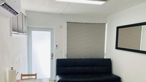 A room with a black sofa, a dining table with chairs, a window with blinds, and an air conditioning unit. - California Apartments (Yabucoa)