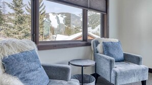 Habitación con dos sillones grises y una pequeña mesa redonda, una ventana con vistas a montañas nevadas y árboles.