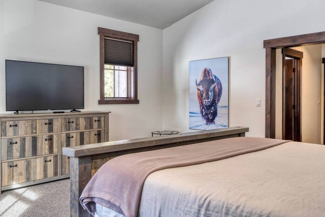 A bedroom with a bed, a TV on a wooden cabinet, a window with blinds, and a painting on the wall.