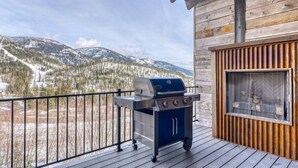 Una terraza de madera con una parrilla y una ventana con vistas a un paisaje montañoso nevado.