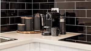 Una cocina con salpicadero de azulejos negros, cafetera, hervidor y jarra de café sobre el mostrador.