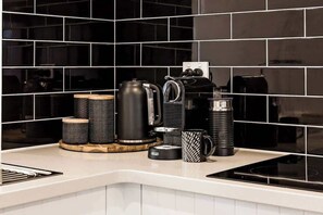 Ett kök med svarta kakelplattor på väggen ovanför arbetsbänken, en kaffebryggare, en vattenkokare och en kaffekanna på bänken.