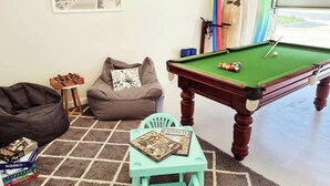Una mesa de billar con superficie de fieltro verde, un sillón de peluche, una pequeña mesa con libros y un soporte para tablas de surf contra la pared.