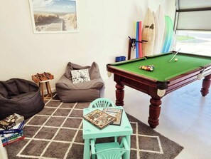 Ett biljardbord med en grön filtduk, en sittpuff, ett litet bord med böcker och ett surfbrädeställ mot väggen.