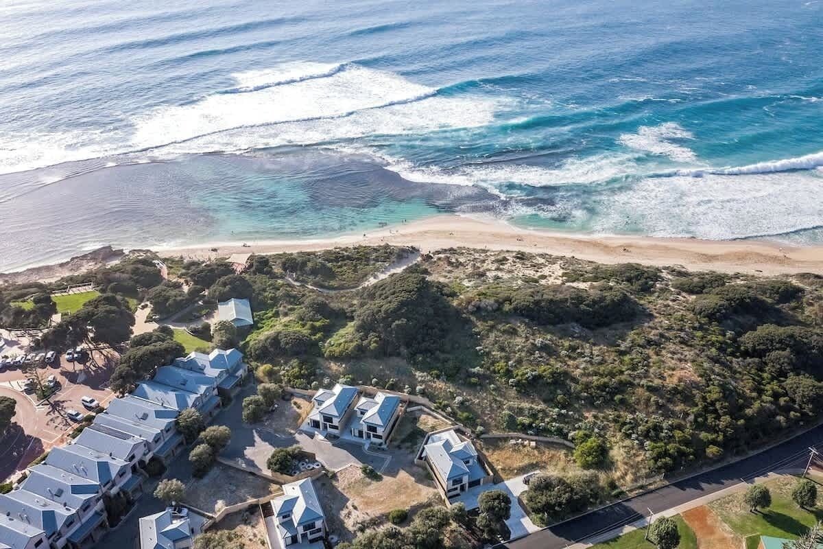 An aerial view of a coastal property with a beach, a parking area, and a row of houses with blue roofs.